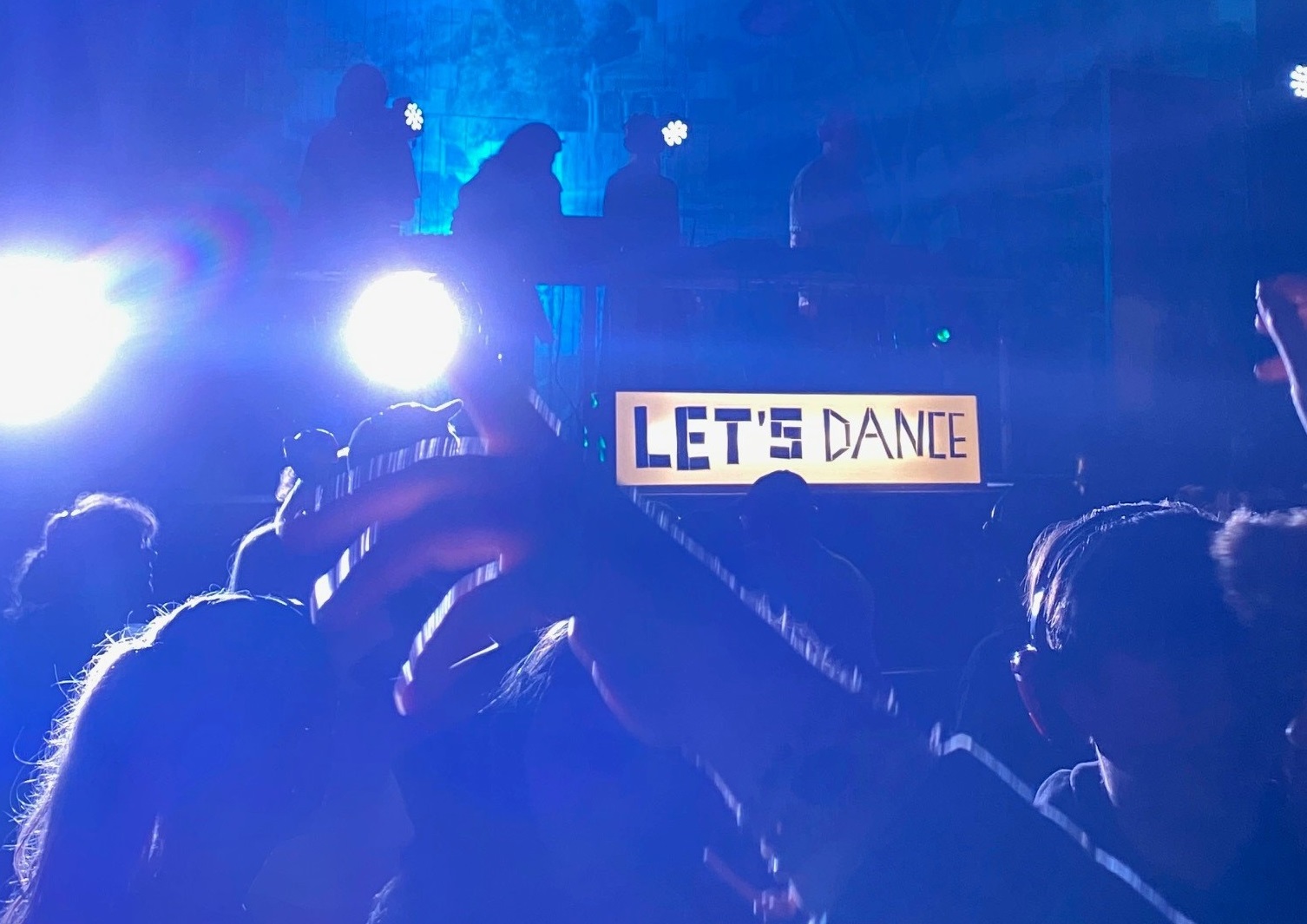Images shows people in silhouette dancing with an illuminated sign that says Let's Dance